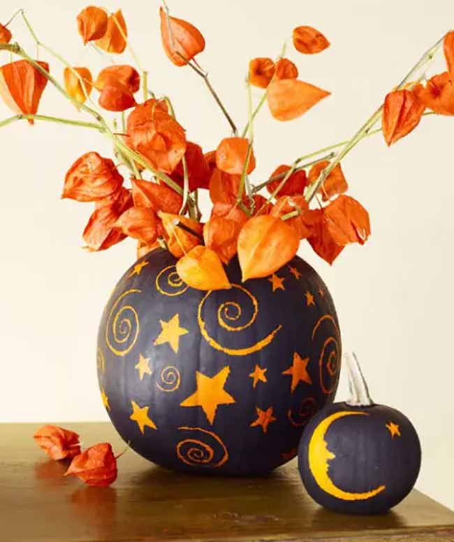 A table centerpiece features a large black pumpkin as a vase, decorated with gold stars and swirls, accompanied by a smaller pumpkin with a crescent moon face.