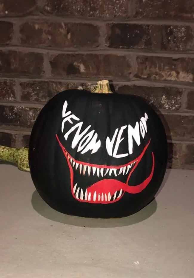 A large black pumpkin is decorated as Marvel’s Venom, featuring a menacing white grin, sharp teeth, a red tongue, and the word “VENOM” in jagged white font.