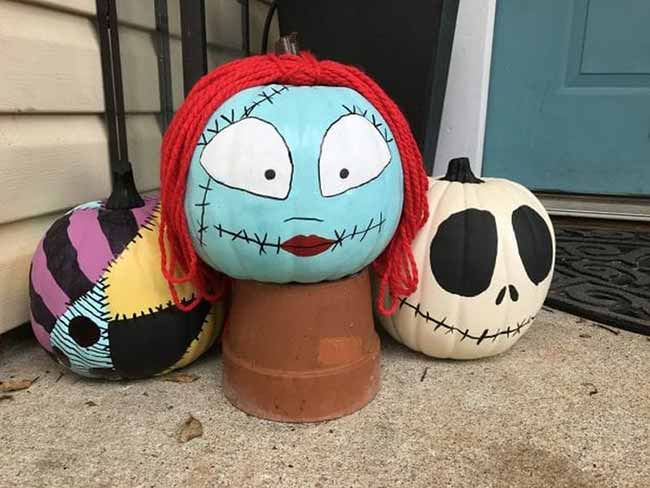 A large pumpkin is painted as Sally from The Nightmare Before Christmas, with a light blue face and red yarn hair, placed next to a matching Jack Skellington pumpkin.