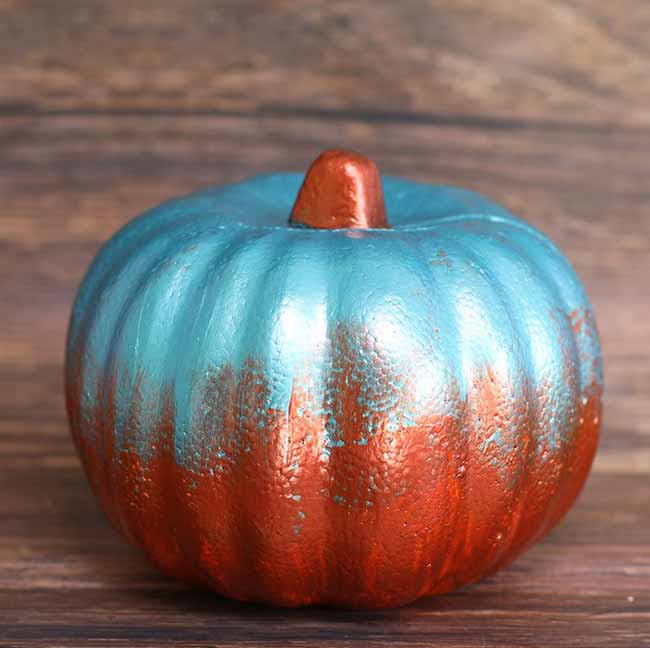 A small round pumpkin features a two-tone finish, with a metallic turquoise top fading into a textured coppery bottom.