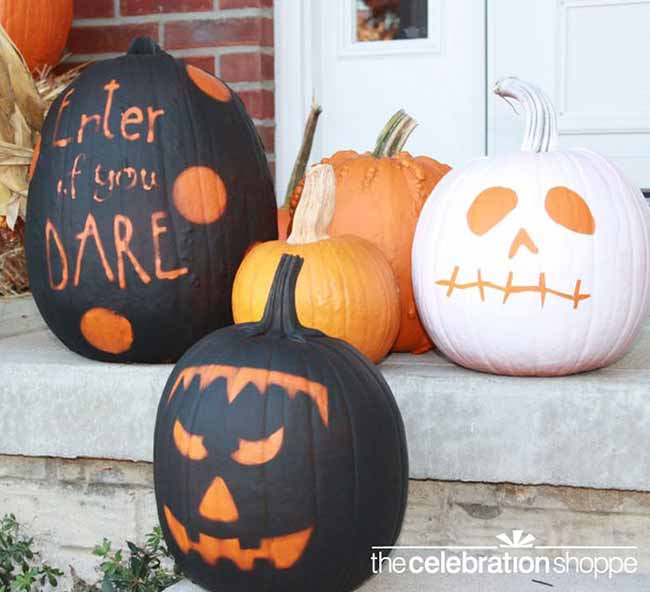 A family of brightly colored pumpkins is stacked on a front porch, including two black pumpkins with orange polka dots and carved phrases, and a white pumpkin with a spooky face.
