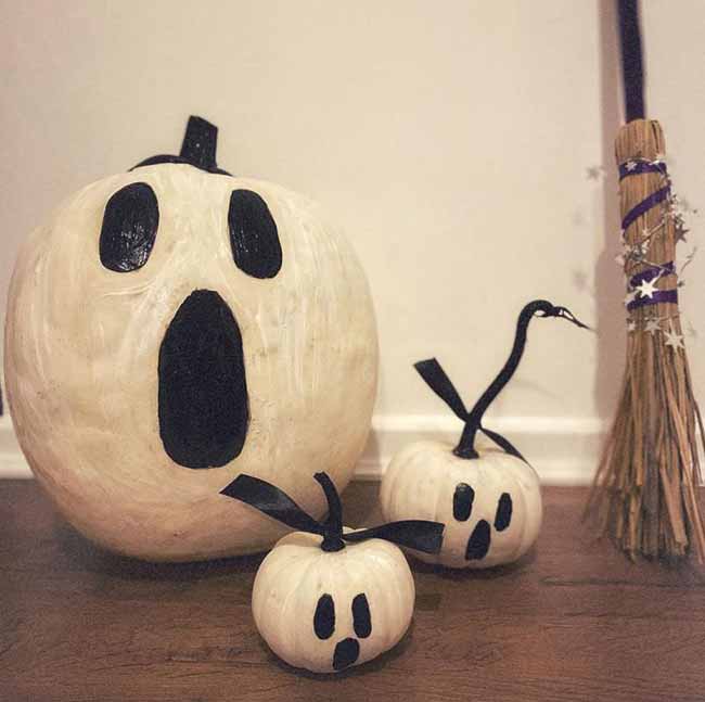 A large white pumpkin with a classic screaming ghost face is joined by two mini ghost pumpkins that have simple black ribbons tied to their stems.