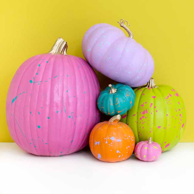 A collection of pumpkins in various sizes is painted in bright colors like neon pink, light purple, and lime green, with splashes of contrasting paint for a fun, modern effect.