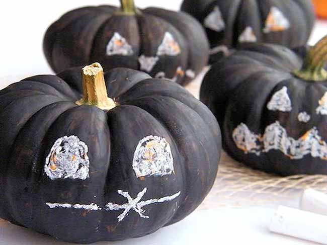 A cluster of small dark pumpkins is decorated with simple spooky faces drawn in white chalk, with slightly smudged details for a casual effect.