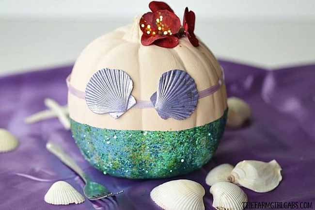 A pumpkin is transformed into a little mermaid, featuring a sparkly teal tail, a purple seashell top, and a few red flowers on its head.