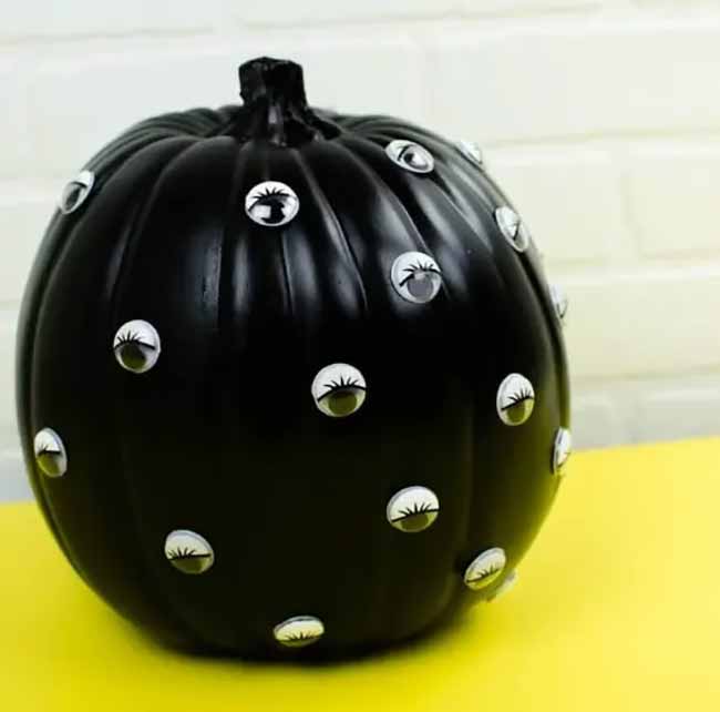 A glossy black pumpkin is covered with numerous small googly eyes, giving it a playful, watchful look.
