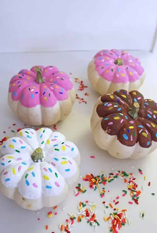 Four small pumpkins are painted to look like iced donuts, each with a different frosting color and colorful sprinkles on top.