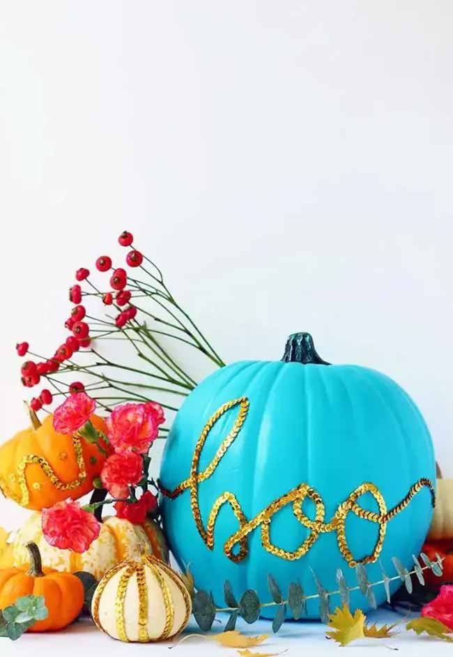 A vibrant turquoise pumpkin is decorated with the word “boo” outlined in gold sequins, surrounded by smaller embellished pumpkins and fall foliage.